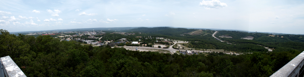 [Four photos stitched together giving a panoramic of green trees in foreground with buildings and road in middstanhe sky.]